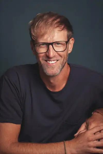 Ein lächelnder Axel Meyer mit Brille und dunklem T-Shirt blickt freundlich in die Kamera, vor einem ruhigen dunklen Hintergrund im Porträt.